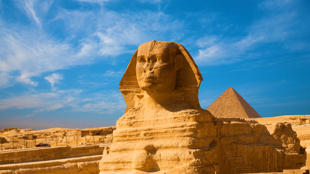 Full length body profile of Great Sphinx including head, feet with great pyramid of Menkaure in background on a clear, blue sky day in Giza, Egypt empty with no people. (photo via pius99 / iStock / Getty Images Plus)