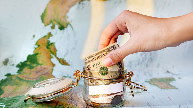 Collecting money for travel. Glass tin as moneybox with cash (Photo via Aksenovko / iStock / Getty Images Plus)