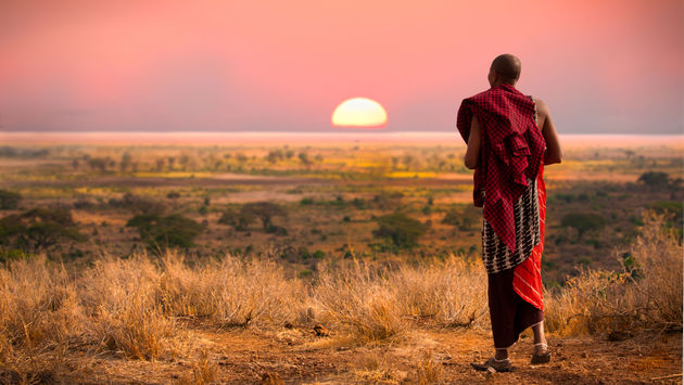 Masai man, wearing traditional blankets, overlooks Serengeti in Tanzania as the colorful sunset fills the sky. Wild grass in the forground. (photo via jocrebbin/iStock/Getty Inages Plus)