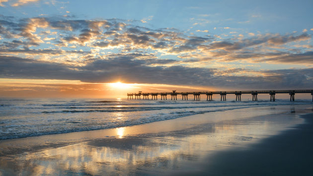Sun rising over horizon and pier, beach illuminated with sunlight, beautiful sky reflected on the beach. Jacksonville Florida, USA. (photo via MargaretW / iStock / Getty Images Plus)